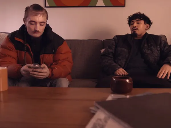 Two friends sitting on a couch, one friend is on his phone.