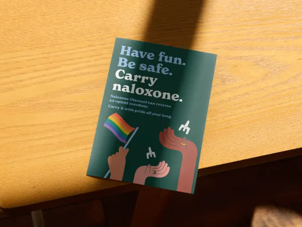 Image of a postcard on a dark background with hands holding a Pride flag and naloxone