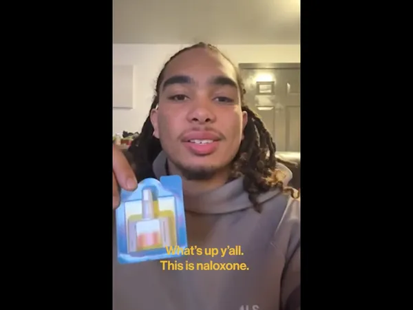 A person holding up a naloxone nasal spray while talking to the camera.