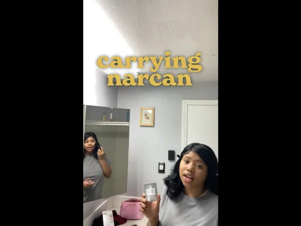 A person doing their make-up in a bathroom, turning to the camera and holding naloxone nasal spray.